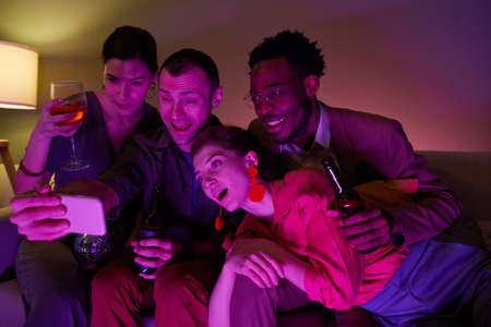 Diverse group of friends taking selfie or video chatting together during indoor party lit by purple...