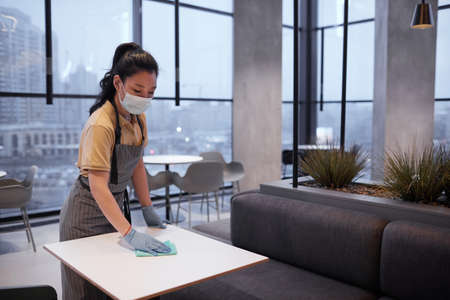 Portrait of young female worker wearing mask while cleaning tables in restaurant or food court...
