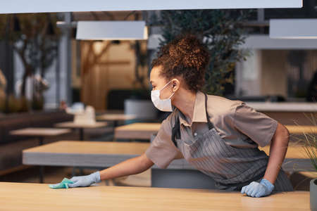 Side view portrait of young woman wearing mask while cleaning tables in modern restaurant interior, safety concept, copy spaceの写真素材
