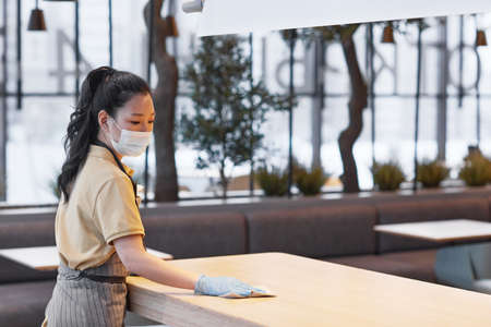 Portrait of young Asian woman wearing mask while cleaning tables in modern restaurant interior, safety concept, copy spaceの写真素材