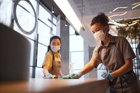 Portrait of two female workers wearing masks while cleaning tables in modern restaurant interior, safety concept, copy spaceの写真素材