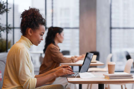 Side view portrait of young African-American woman using laptop while working at table in cafe, copy spaceの写真素材