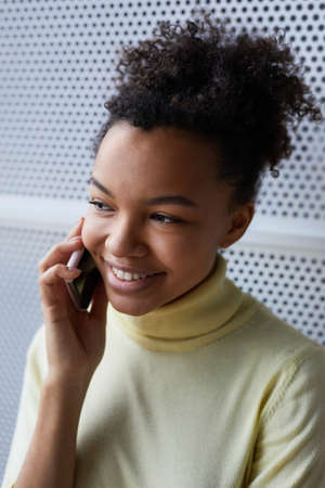 Vertical close up portrait of young African-American woman speaking by smartphone and smiling cheerfullyの写真素材