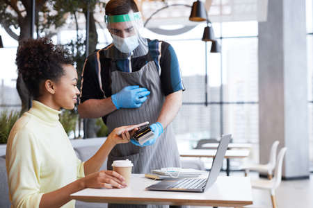 Portrait of waiter wearing face shield and gloves while serving customer in cafe with safety measures, copy spaceの写真素材
