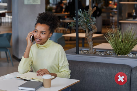 Portrait of young African-American woman speaking by smartphone while studying or working at cafe table with social distancing, copy spaceの写真素材