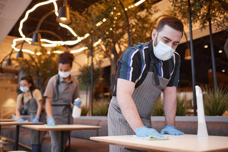 Low angle view at workers wearing masks in row while cleaning tables in cafe interior, covid safety concept, copy spaceの写真素材
