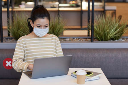Portrait of Asian young woman wearing mask while working at table in cafe with social distancing...