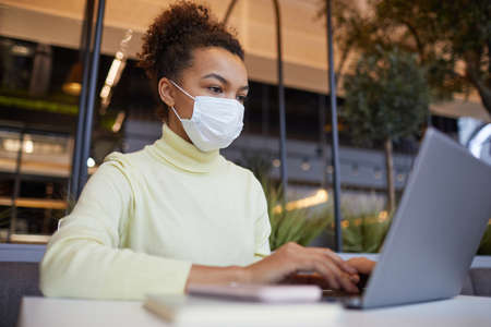 Low angle portrait of Asian young woman wearing mask and using laptop while working at table in cafe