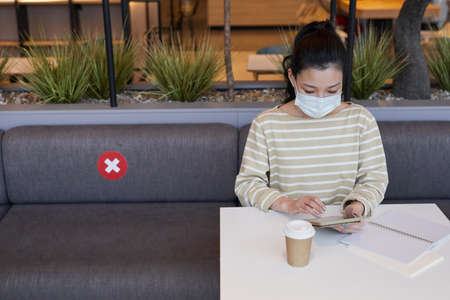 Portrait of Asian young woman wearing mask and using smartphone while working at table in cafe