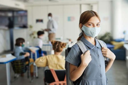 Waist up portrait of cute girl wearing mask in school classroom and looking at camera , safety...