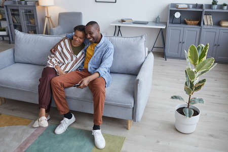 High angle portrait of happy African-American couple watching TV or movies together while sitting on...