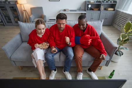 High angle view at group of sports fans wearing red while watching game match at home and sitting on...