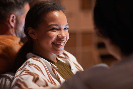 Portrait of young African-American woman smiling happily while watching TV with friends at home lit...