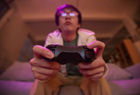 Low angle view at Asian young man holding gamepad and playing video games at home lit by purple light, copy spaceの写真素材
