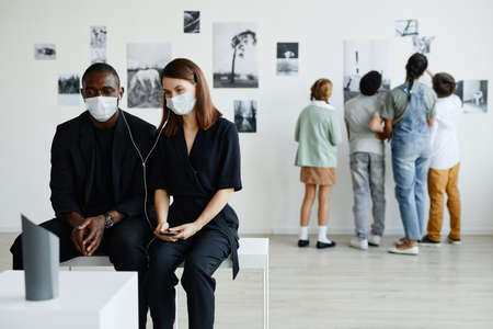 Minimal portrait of mixed-race couple in art gallery wearing masks and listening to audio guide, copy spaceの写真素材
