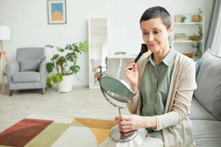 Portrait of smiling mature woman doing makeup and looking in mirror in cozy home setting, copy spaceの写真素材