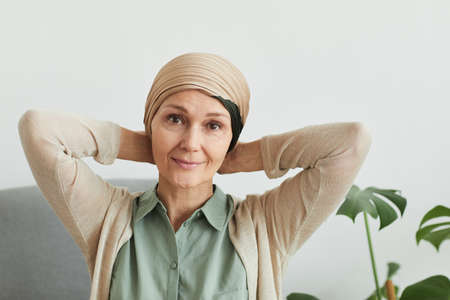 Portrait of mature woman looking at camera while putting on headscarf at home and getting ready to...