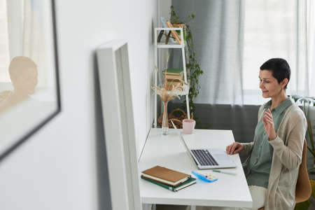 Side view at smiling mature woman waving hello during video call via laptop in cozy home setting,...