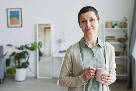 Waist up portrait of mature woman smiling at camera while standing by window and holding cup of...