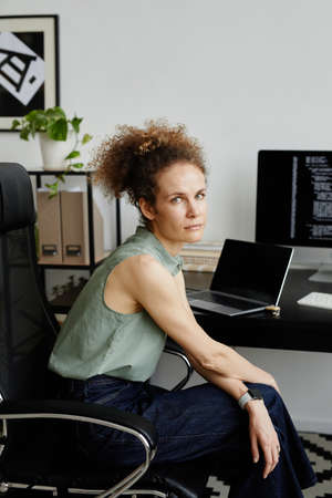 Portrait of young woman sitting on chair and looking at camera while worknig at her workplace at officeの写真素材