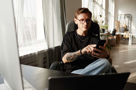 Young businessman sitting on chair and typing a message on mobile phone while working at the table at officeの写真素材