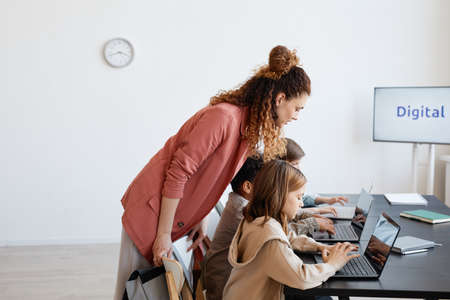 Side view portrait of female teacher helping children using laptop during IT class in school, copy...