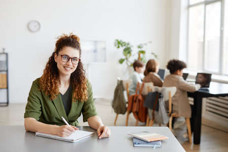 Portrait of young female teacher smiling at camera with diverse group of children in background,...