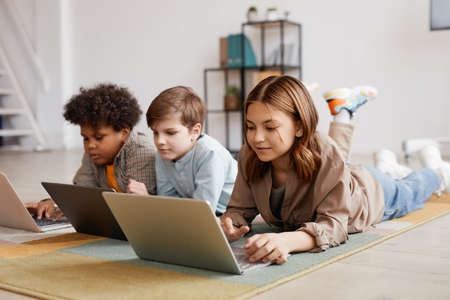 Full length view at group of three kids using computer devices while laying on floor, focus on smiling girl in foregroundの写真素材