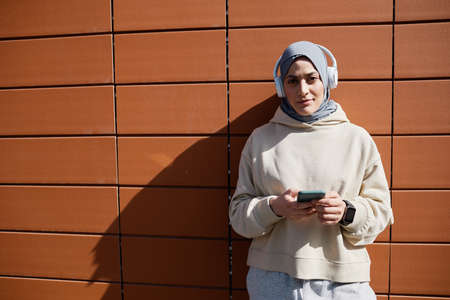 Graphic waist up portrait of contemporary Middle-Eastern woman using smartphone in city lit by sunlight and wearing headphones, copy spaceの写真素材