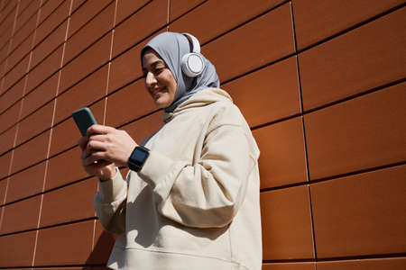 Low angle portrait of contemporary Middle-Eastern woman using smartphone in city lit by sunlight and wearing headphones, copy spaceの写真素材