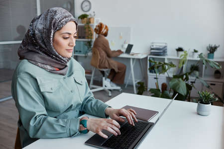 Portrait of modern Middle-Eastern woman wearing headscarf in office and using laptop, copy spaceの写真素材