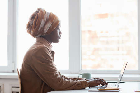 Side view portrait of young African-American woman wearing headscarf while sitting against window in office and using computer, copy spaceの写真素材