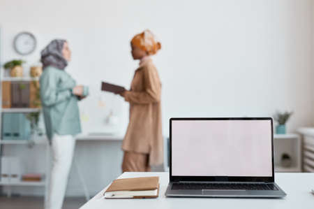 Close up of open laptop with blank white screen with two ethnic women working in background, copy spaceの写真素材