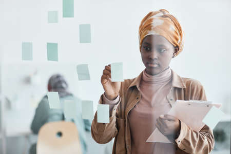 Waist up portrait of modern African-American woman working in office and placing sticker notes on...