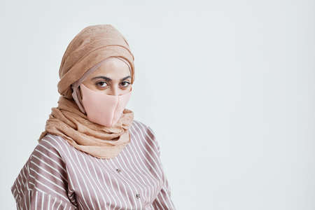 Portrait of modern Middle-Eastern woman wearing mask and looking at camera while posing against white wall, copy spaceの写真素材