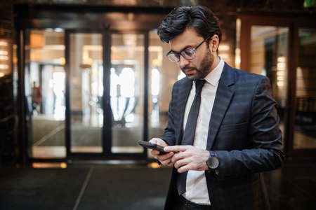 Waist up portrait of successful Middle-Eastern businessman using smartphone while standing in hotel...