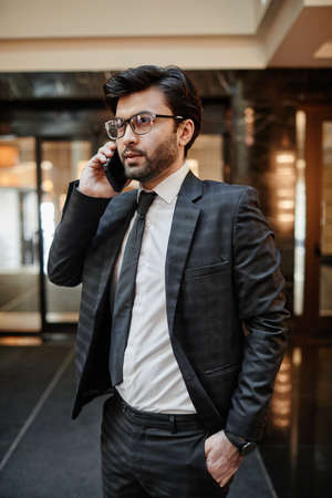 Vertical waist up portrait of successful Middle-Eastern businessman speaking by smartphone while standing in hotel lobbyの写真素材