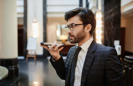 Side view portrait of successful businessman recording voice message to smartphone while standing in hotel lobby, copy spaceの写真素材