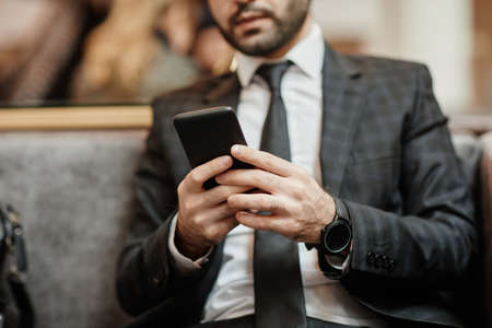 Cropped shot of successful businessman using smartphone while sitting in armchair at hotel lobby, copy spaceの写真素材