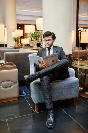 Vertical full length portrait of successful Middle-Eastern businessman reading documents while sitting in armchair at hotel lobbyの写真素材