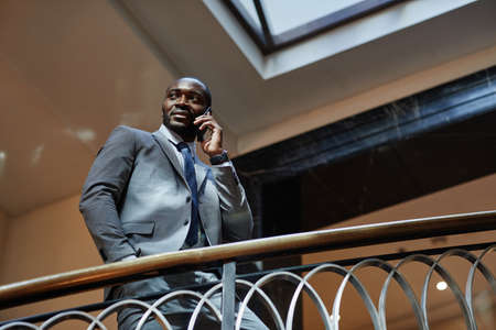 Low angle portrait of successful African-American businessman speaking by smartphone while standing...