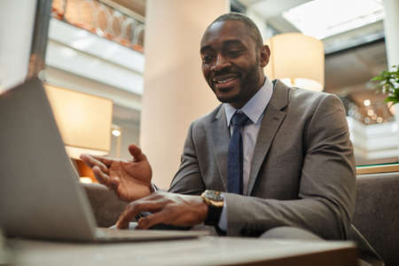 Portrait of successful African-American businessman using laptop during online meeting in hotel lobby, copy spaceの写真素材
