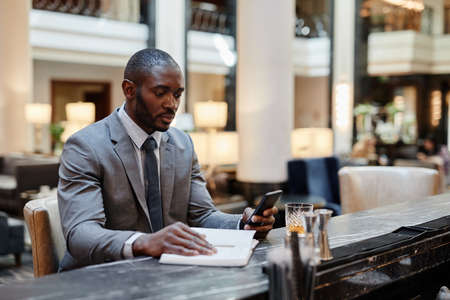 Portrait of successful African-American businessman using smartphone while relaxing at bar in hotel lobby, copy spaceの写真素材