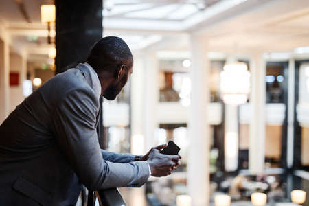 Side view portrait of African-American businessman standing at balcony over luxurious hotel lobby,...