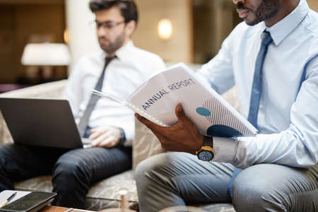 Cropped portrait of unrecognizable businessman reading report while working in hotel lounge, copy...
