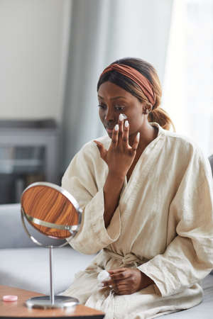 Vertical portrait of young African-American woman using face cream or moisturizer, skincare and beauty routine conceptの写真素材