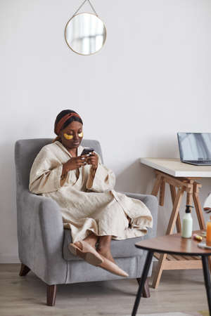Minimal full length portrait of young African-American woman enjoying skincare routine at home and using smartphone, copy spaceの写真素材