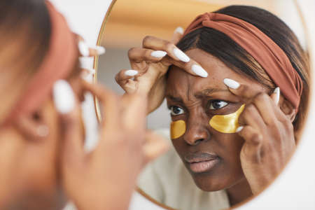 Portrait of young African-American woman doing skincare routine at home and popping zit while looking at mirror, copy spaceの写真素材