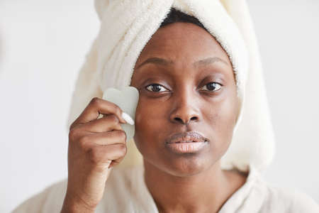 Front view portrait of beautiful African-American woman doing face massage while enjoying skincare routine at home and looking at camera, copy spaceの写真素材