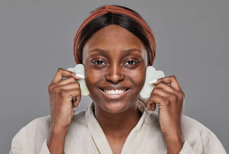 Front view at young African-American woman enjoying face massage with gua sha stones and smiling at...